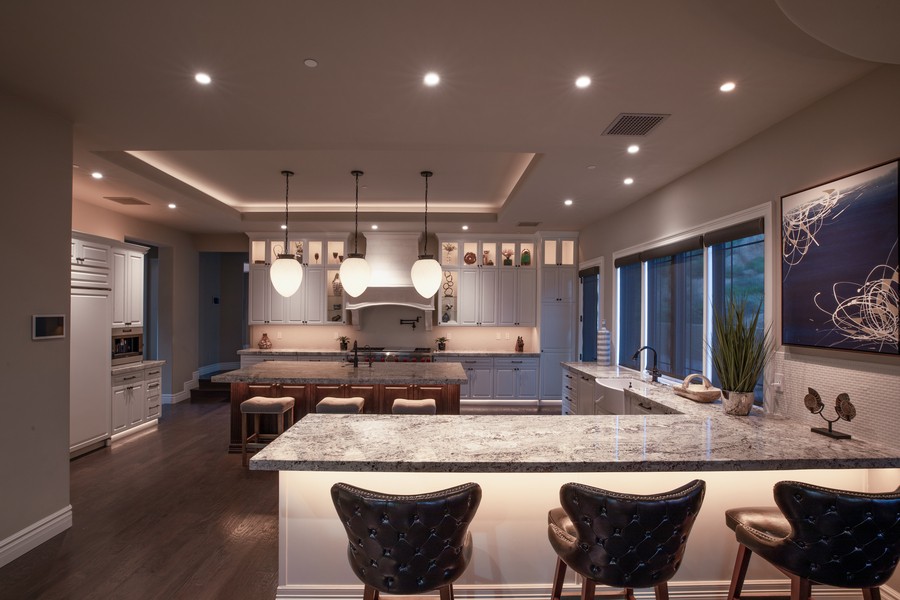 A spacious, modern kitchen with gray cabinets, granite countertops, and a large center island with a bar area. It is illuminated by different shades of lighting.