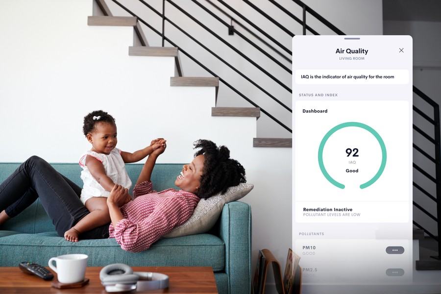 A woman lying on a sofa playfully playing with her child on her stomach, both smiling and looking at each other. To the right, an overlay of a smart home interface displaying 'Air Quality' data for the living room.