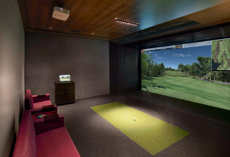 A home golf simulator room featuring a large screen display, artificial turf mat for golfing, overhead projector, and comfortable seating