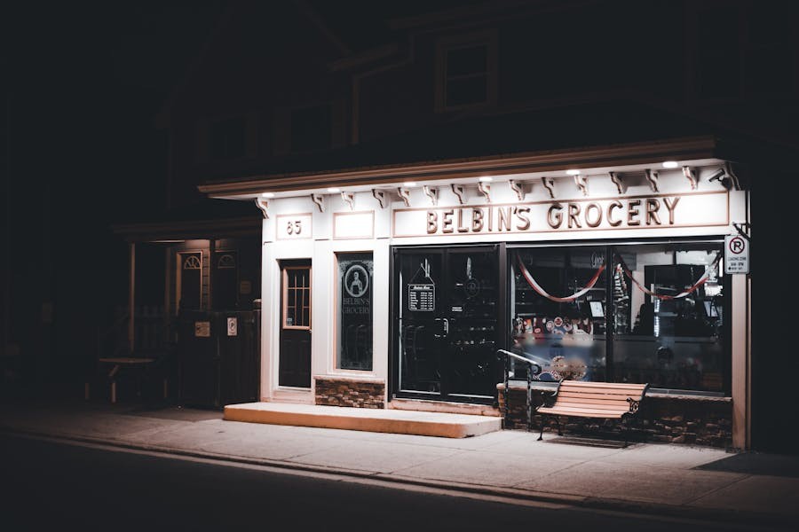 Small storefront labeled "Belbin's Grocery" illuminated at night with exterior lighting, featuring large front windows and a bench on the sidewalk.