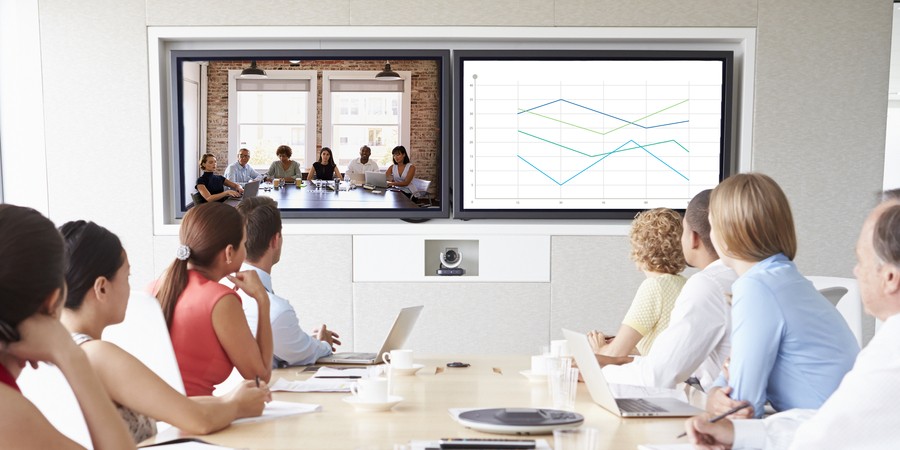 A conference room with a table full of people looking at two screens on the wall–one featuring a video conference and the other a presentation. 