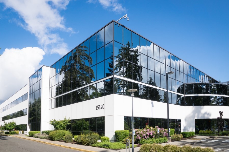 A white office building with lots of windows. There are bushes around the building and the parking lot.